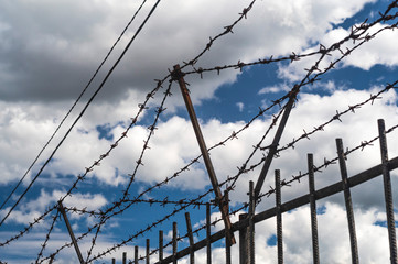 Tall metal fence with barbed wire against a blue sky with clouds