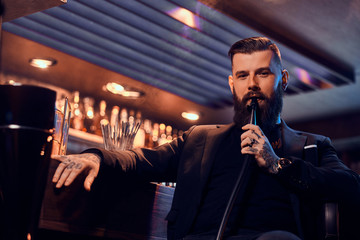 Attractive groomed man with beard and tattooes is smoking hookah while sitting near bar counter.