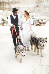 Beautiful hippies groom and bride celebrate their wedding in the desert. Mixed races couple of lovers are standing with dogs in the desert against the background of the decorated space