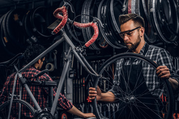 Focused attractive man in glasses is chainging wheel for bicycle at busy workshop.
