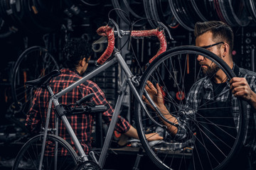 Focused attractive man in glasses is chainging wheel for bicycle at busy workshop.