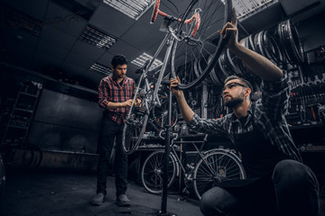 Fototapeta na wymiar Two attractive mans are working on bicycle fixing in dark workshop.
