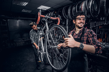 Two attractive repairmans are fixing broken bicycle at their dark workshop.