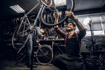 Two attractive repairmans are fixing broken bicycle at their dark workshop.