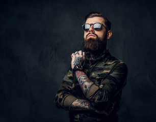 Bearded tattooed guy in military shirt and sunglasses posing with thoughtful look. Studio photo against a dark wall