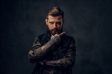 A crazy bearded man in the military shirt and denim vest with tattooed hands showing the fuck sign. Studio photo against a dark wall