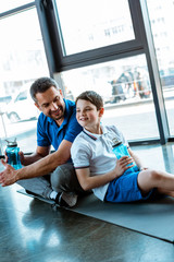 happy father and son sitting on fitness mat with sport bottles at gym