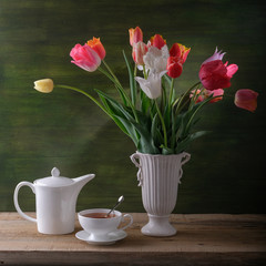 A cup of tea, a white kettle and tulips in a vase on the table.