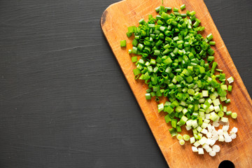 Chopped green onions on a rustic wooden board on a black surface, overhead view. From above, flat lay, top view. Copy space.