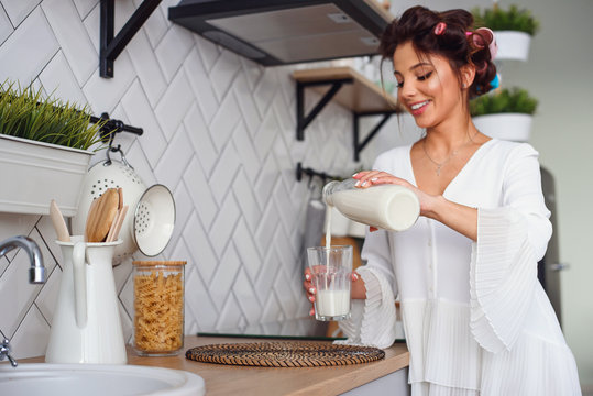 Beautiful smiling woman pours fresh milk from a carafe into a glass, in the stylish cozy kitchen at the morning. Healthy eating and diet concept. - Powered by Adobe