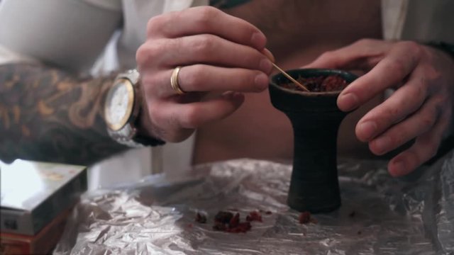 Married Caucasian man in a white unbuttoned shirt with a tattoo with watch and ring sprinkle and packed red tobacco in a clay bowl using a toothpick for smoking hookah close-up.