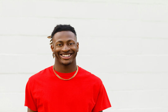 African American Teen Male wearing a red t-shirt and jeans Smiling against a white painted brick wall