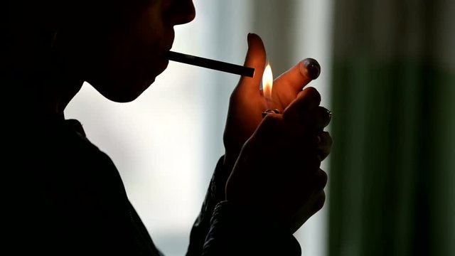 Close-up of the silhouette of a woman's face sticking a cigarette into her lips and igniting her with a lighter. The grim concept of the harm of smoking in a low key