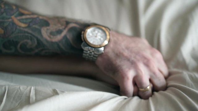 Married couple in love holding hands tight, close up. Man with tattoos, watch and ring holds woman hand, selective focus. Male and female hands crossed on white cotton sheets in bed. Sex concept. 