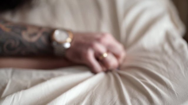 Married couple in love holding hands tight, close up. Man with tattoos, watch and ring holds woman hand, selective focus. Male and female hands crossed on white cotton sheets in bed. Sex concept. 