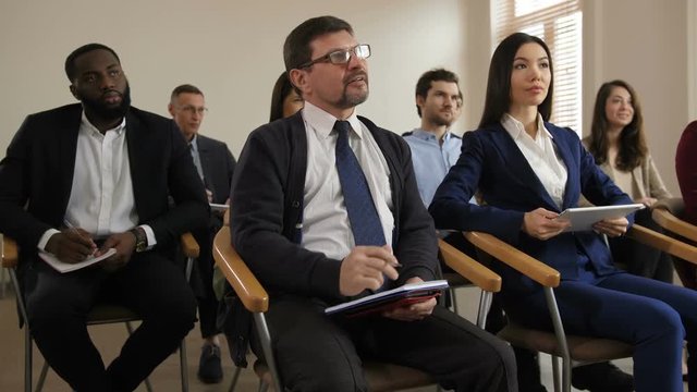 Attentive multinational business people writing notes while coach giving speech. Closeup spectacled middle aged man making clarifying question to speaker during corporate business training