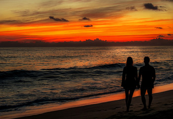 Man and woman couple in love holding hands during red sunset at the beach with waves at the ocean in Mexico
