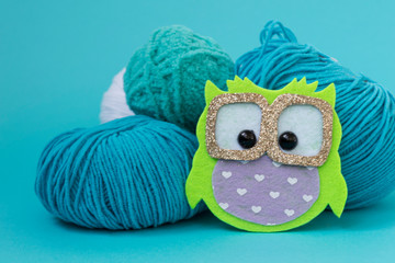 Self-made soft toy owl made of felt, on a color monophonic background. The owl stands next to the balls of yarn. There is a place for text.
