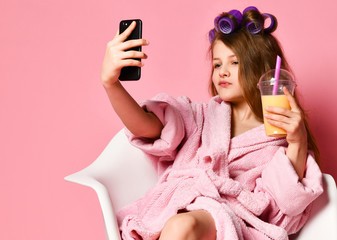 Young cheeky lady teen girl in bathrobe and with hair in curlers drinks smoothie fresh juice and takes cool fashion look selfie on pink