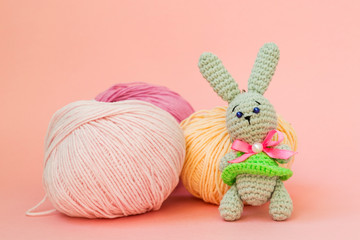 Gray knitted hare stands on the background of multi-colored balls of yarn on a pink background. There is a place for text.