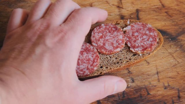 A man's hand takes a sandwich with sliced meat salami for a snack or for breakfast. A slice of black bread with mugs of dry sausage for breakfast on a wooden board.