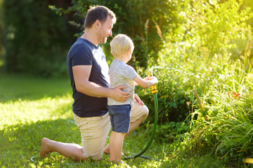 Funny little boy with his father playing with garden hose in sunny backyard. Preschooler child...