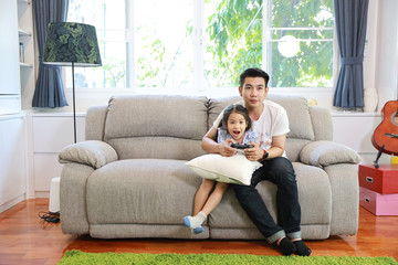Obraz premium Happy Asian family, father and daughter are holding joystick and playing video game on the sofa in the living room with happy smiling face (relaxation and technology concept)