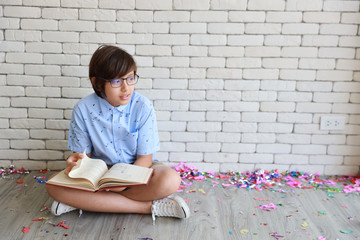 Fototapeta na wymiar cute and smart little boy in blue shirt with glasses sitting and reding red books with intended face and white wall background
