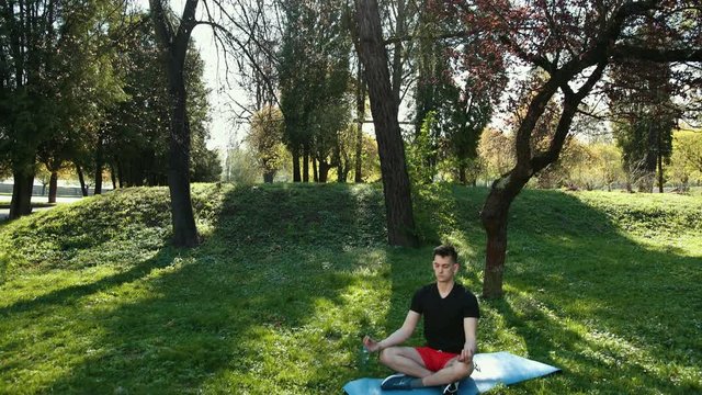 Young Man Practicing Yoga Outdoors At Park Sitting On Mat In Lotus Pose Closed Eyes Meditating. amera movement from tree to person. Dressed In Arable T-Shirt, Red Shorts Shut Down Eyes. Slow Motion