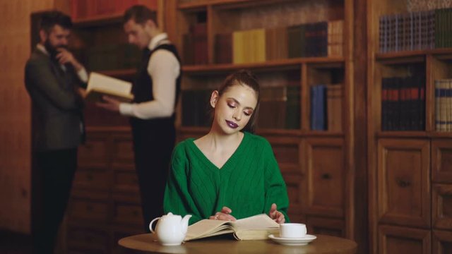 Woman in library read book and drinking coffee from cup. Literature cafe with cute girl and men's on background. Student life in university. Concept of reading book and study poetry.