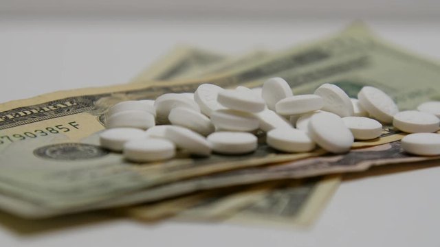 Pill bottle pours out white pills onto stacks of money.