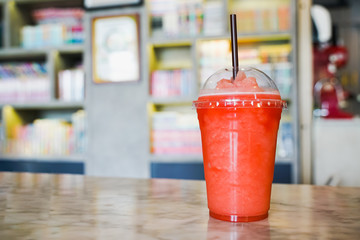 fruit frappe in transparent plastic glass and brown straw on table with blurred bookshelf