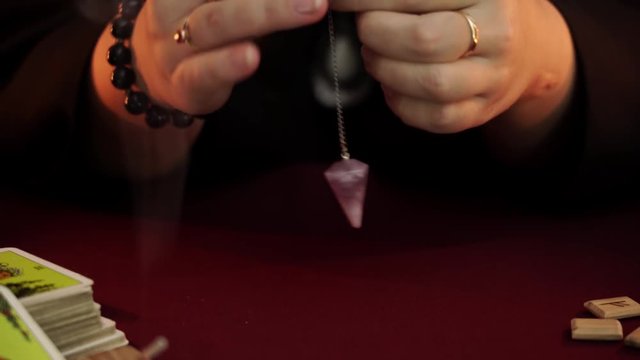 A witch is fortune teller in robe performs a rite of black magic. Pendulum say No. Magical ritual. Tarot cards, white candles and runes. Occult, esoteric and divination concept. Mystic background.