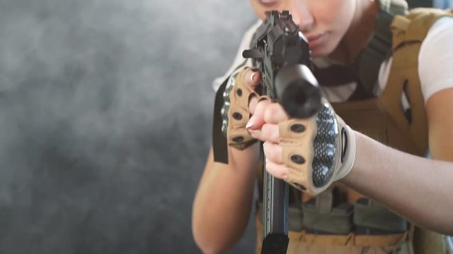 Airsoft female shooter playing strikeball, sets her sights on enemy with automatic assault rifle. Studio shot, dark smoky background.