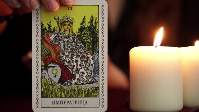 A witch is fortune teller in mantle shows tarot card. White candles on table. Occult, esoteric, divination and wicca concept. Translation: The Empress.
