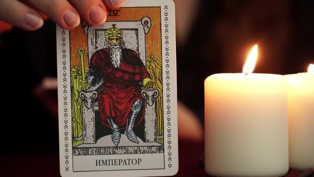 A witch is fortune teller in mantle shows tarot card. White candles on table. Occult, esoteric, divination and wicca concept. Translation: The Emperor.