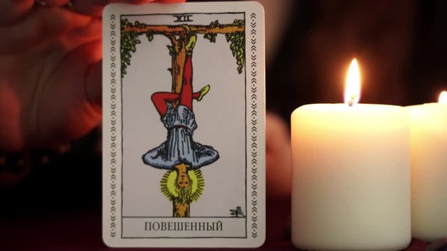 A witch is fortune teller in mantle shows tarot card. White candles on table. Occult, esoteric, divination and wicca concept. Translation: The Hanged Man.