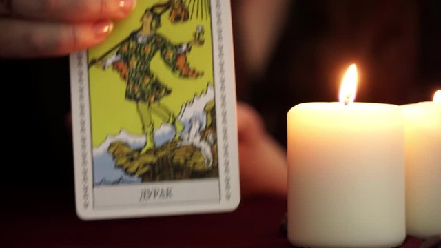 A witch is fortune teller in mantle shows tarot card. White candles on table. Occult, esoteric, divination and wicca concept. Translation: The Fool.