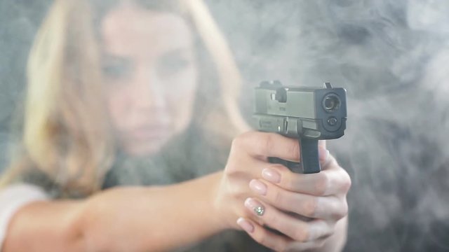 Armed beautiful blonde woman wearing protective headphones and plate carrier, shoots with gun at a target in the darkness with smoke clouds