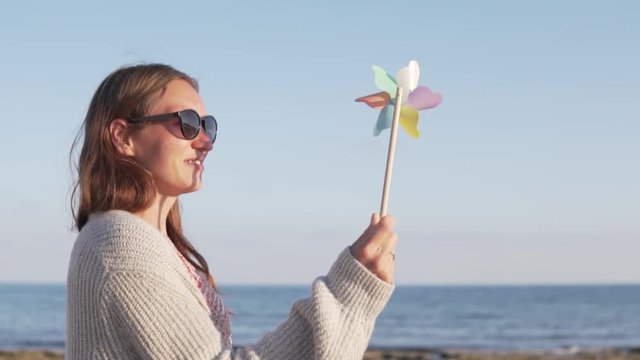 Beautiful woman in sunglasses on summer holiday playing with toy windmill. A woman stands on the beach, she is holding a toy tiller in her hands. The colored blades swirl beautifully in the wind