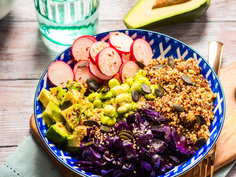 Buddha bowl with rainbow colors ingredients - avocado, fermented red cabbage, quinoa, radish, green beans, sesame and pumpkin seeds. Dish served on wooden board. Healthy vegan plant based lunch.