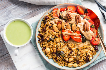 Delicious amaranth porridge topped with home made granola, strawberries and almond butter. Vegan plant based breakfast served with matcha latte on white wooden board. Healthy colorful food concept