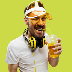 Half-length close up portrait of young man in shirt on yellow background. Male model with headphones and drink. The human emotions, facial expression, summer, weekend concept. Smiling and drinking.