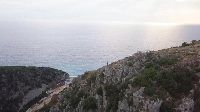 Human standing on a huge cliff looking into the ocean at sunset. Flying drone revealing the scale of Gjipe Canyon and the Adriatic Sea.