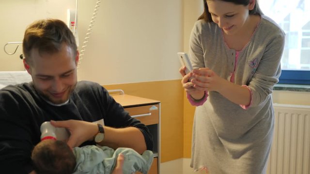 Young mother take phone picture of father first time feeding his newborn baby with a milk bottle nutrition visiting them in maternity hospital couple days after birth