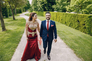 Fototapeta premium Beautiful woman in a long red dress walks around the city with her husband in a blue suit and with a beard