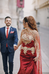 Beautiful woman in a long red dress walks around the city with her husband in a blue suit and with a beard