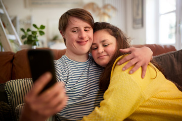 Loving Young Downs Syndrome Couple Sitting On Sofa Using Mobile Phone To Take Selfie At Home