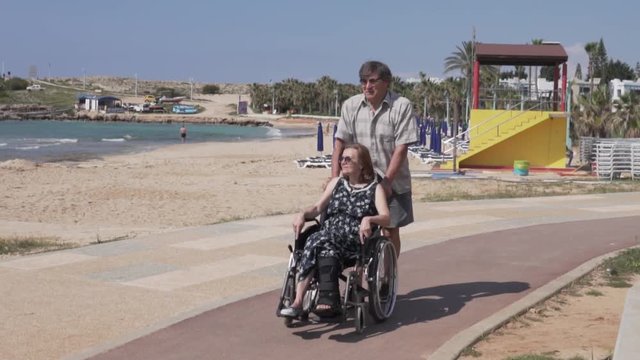 An elderly man carries a woman in a wheelchair along the sea. The husband carries his wife with a broken leg in the wheelchair past the window. A woman with a broken leg in an orthosis in a wheelchair