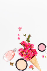 Beauty background. Waffle cone with pink spring flowers and different makeup cosmetic on light background. Face powder lipstick concealer bottle of perfume makeup brush. Flat lay top view copy space.